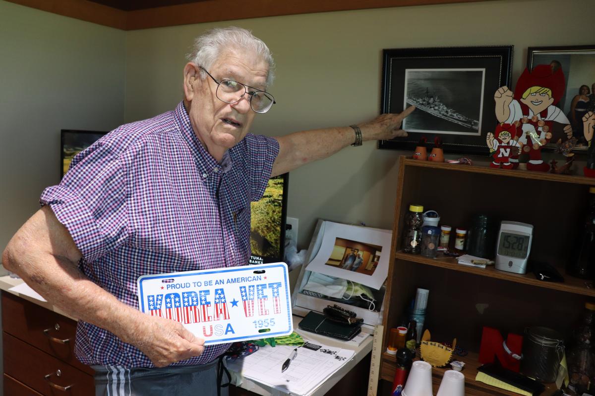 Kyle Lantz with Korean War Veteran license plate pointing at photo of ship